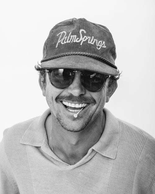 Justin Eldridge smiling with a golf pick in his mouth and wearing a "Palm Springs" hat and sunglasses