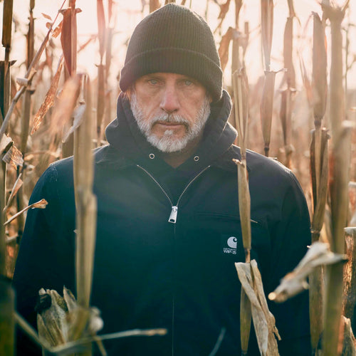 Steven Lippman wearing a black Carhartt jacket and beanie standing in a field with tall grass