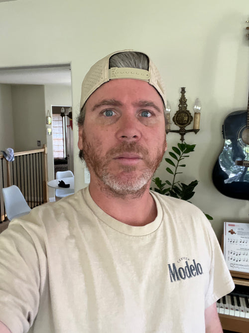 Val  Arkenstone wearing a beige cap and 'Modelo' t-shirt in a room with a guitar and plant