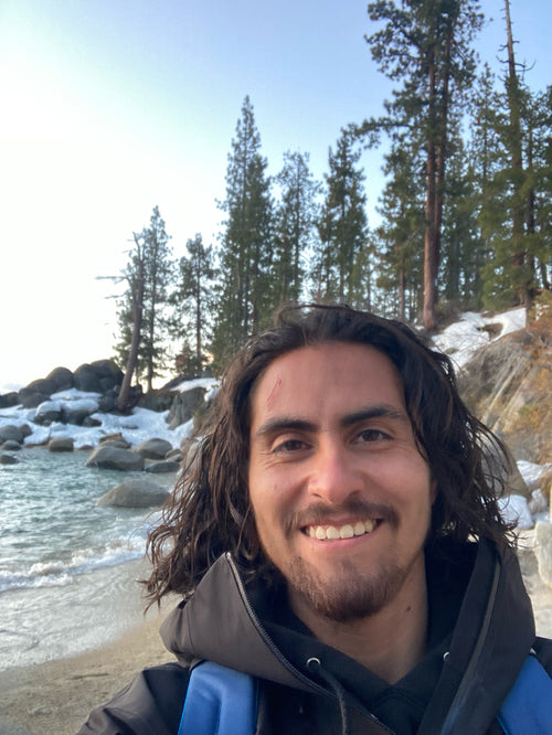 Chris Rivera standing on a beach with water, rocks, snow, and trees in the background