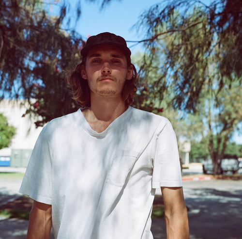 Conner Frost standing in front of some trees wearing a white shirt and black hat