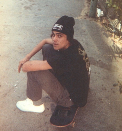 Derricke Nua sitting on a skateboard wearing a black beanie and black shirt with a graphic design