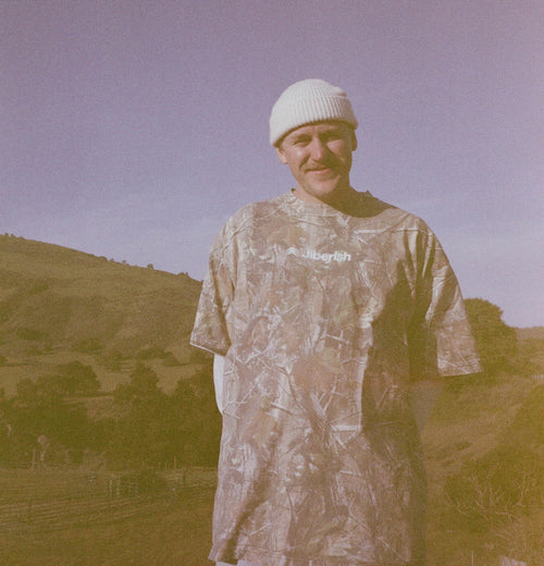 Will Mault wearing a camouflage shirt and white beanie standing in a field with hills in the background