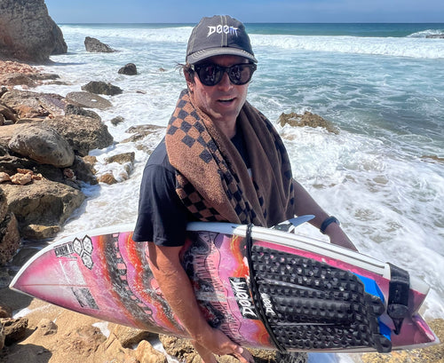 Dillon Perillo holding a colorful surfboard on a rocky beach with ocean waves in the background wearing a black "DOOM" hat