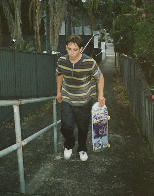 Alex Midler holding a skateboard in an outdoor setting with a fence and trees in the background