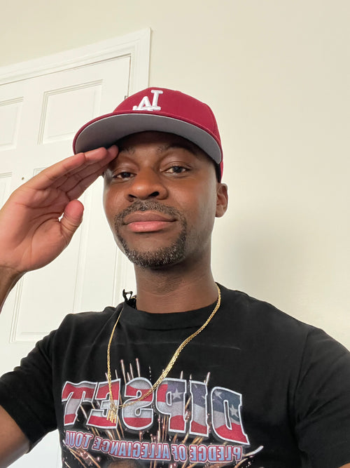 Henri Yoro wearing a red and gray cap with a "LA" logo, black t-shirt and gold necklace indoors.