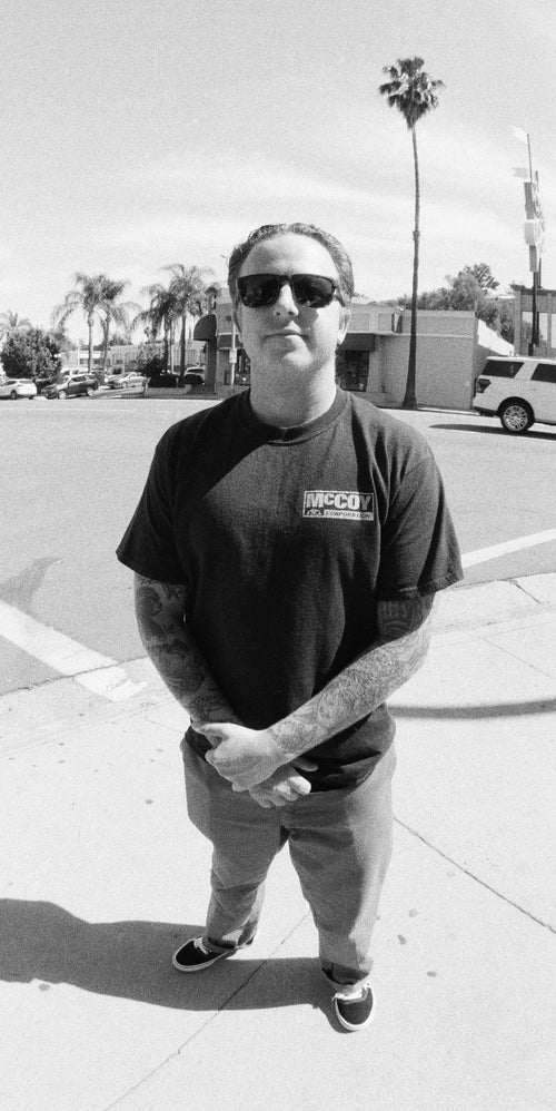 Shane Brown wearing sunglasses and a black t-shirt, standing on a sidewalk with palm trees in the background.