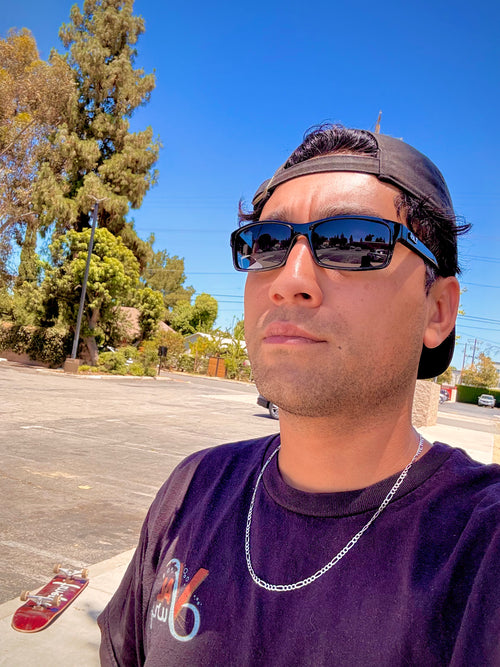 Lalo Trejo wearing sunglasses, a "Val Surf" t-shirt,  and a cap with a clear blue sky and trees in the background