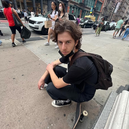 Max Klein sitting on his skateboard while people walk the sidewalk behind him