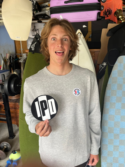Sebastian Mendes sticking his tongue out in front of his surfboard quiver while holding an IPD sticker