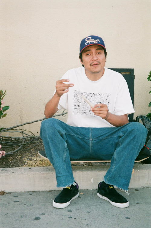 Stevie Perez sitting on a skateboard on a curb wearing a scorpion hat