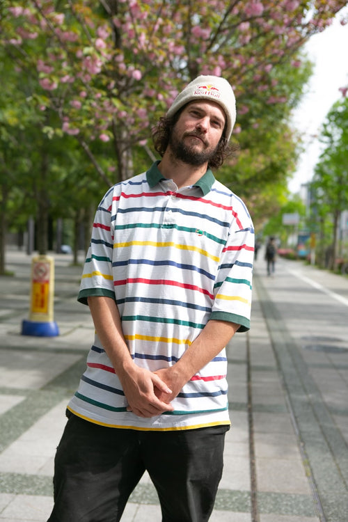 Torey Pudwill standing on a sidewalk wearing a "Red Bull" beanie and a striped polo shirt