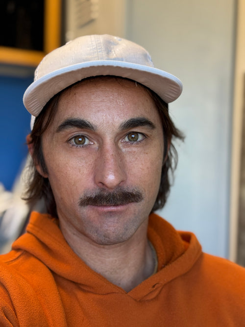 Gabe Taylor wearing a white cap and orange hoodie indoors with a proper mustache