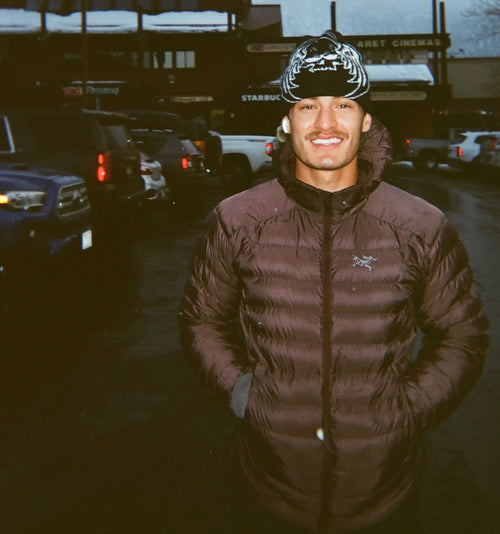 Peter Molina wearing a brown Arcteryx puffer jacket and a black beanie standing in a parking lot with vehicles and a building in the background