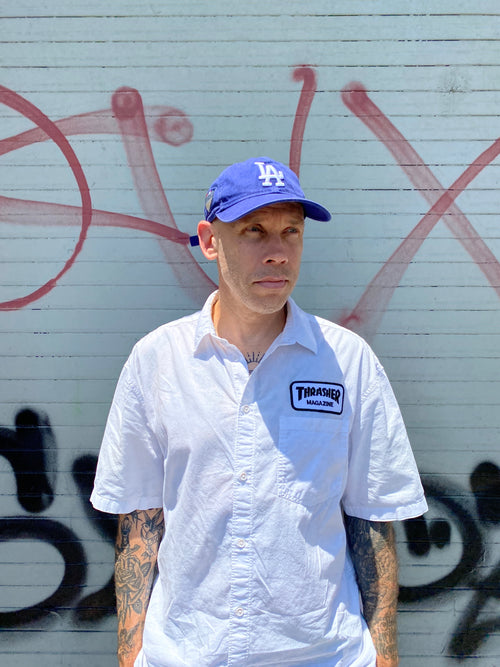 Shane Heyl wearing a white "Thrasher" jacket and blue "LA" cap standing on a graffiti-covered wall